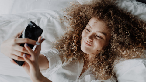 Junge Frau, die lächelnd im Bett liegt und ein Selfie macht
