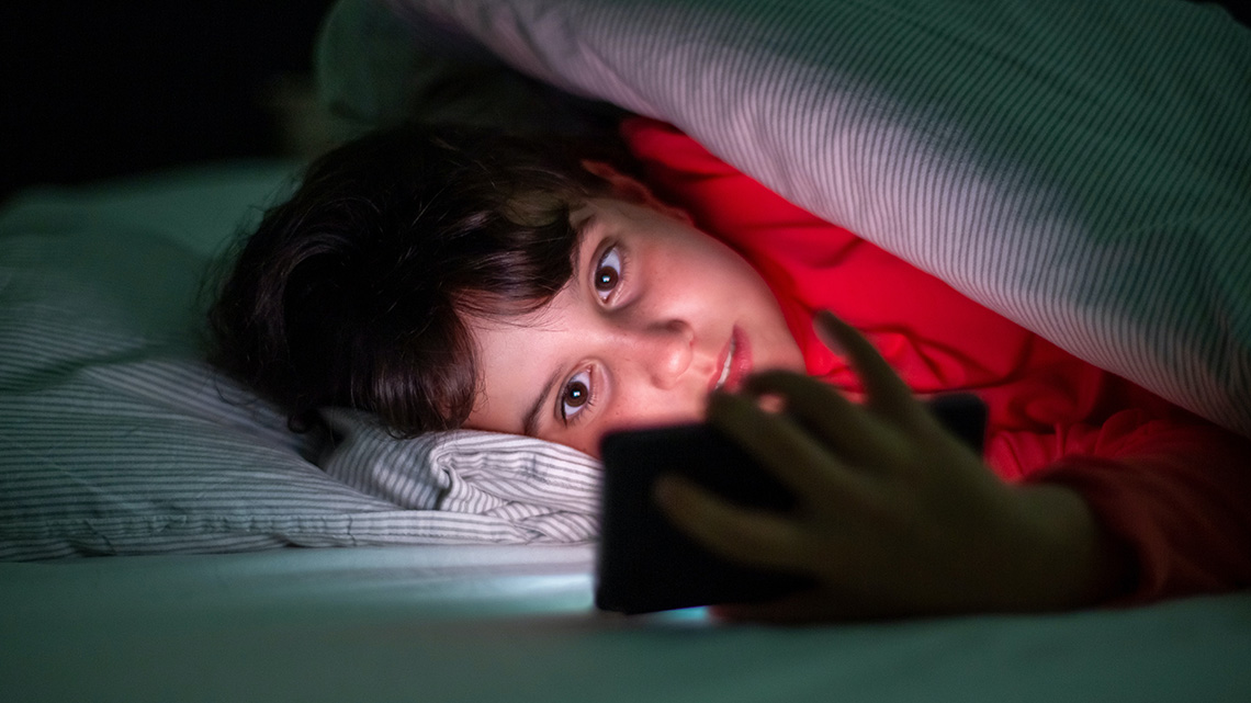 Kind, das auf sein Smartphone schaut und sich unter seiner Bettdecke versteckt